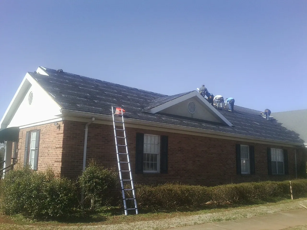 Crew tearing shingles off a brick ranch roof with a ladder set — Emergency Roof Repair work by our crew in Highland