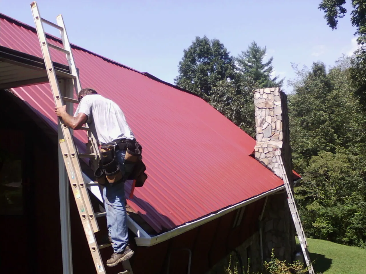 Roofer on a ladder beside a red metal roof and stone chimney — Soffit & Fascia Repair by Roofing Brothers in Highland